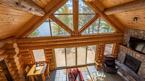 Interior de uma cabana de tronco com área de jantar, lareira e grandes janelas com vista para um lago.