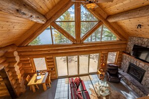 Interior de una cabaña de troncos con comedor, chimenea y grandes ventanales con vistas a un lago.