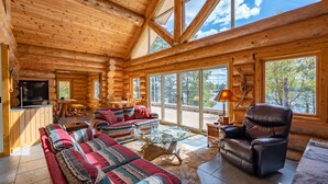 Interior de uma cabana de tronco com uma grande porta de vidro que dá para um deck, um conjunto de sofá, uma poltrona de couro e uma lareira.