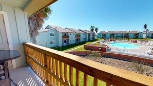 A residential building with a swimming pool, a balcony with a table and chairs, and a clear blue sky. - 2nd Floor, Shared Pool and Hot Tub, Golf Cart Access (Port Aransas)