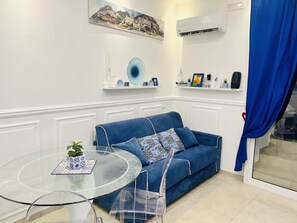 A modern living room with a blue sofa, a glass coffee table, and a clear chair. There is a painting on the wall and a shelf with decorative items.