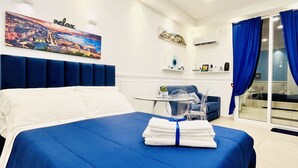 Een moderne hotelkamer met een groot bed, een stadsgezicht schilderij, een klein tafeltje met een stoel, en een schuifdeur met blauwe gordijnen.