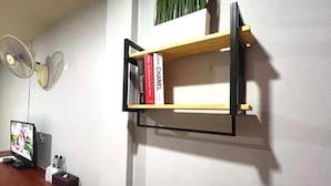 Een kamer met een bureau, een computerscherm, een ventilator en een wandplank met boeken en een plant.