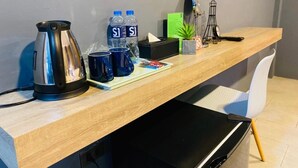 Een moderne hotelkamer met een houten bureau, een waterkoker, twee waterflessen, twee mokken, een kleine plant, een tijdschrift, een afstandsbediening en een zwart elektronisch apparaat.