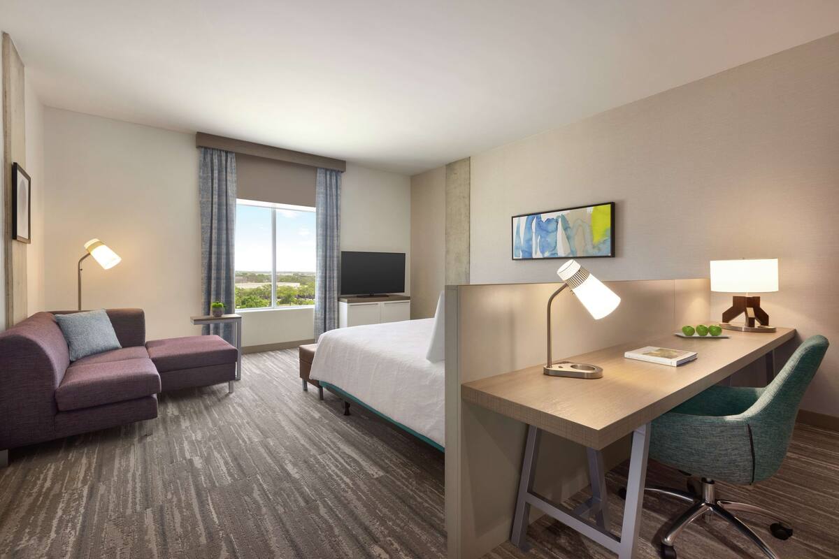 A hotel room with a bed, a desk with a chair, a sofa, a TV, and a window with curtains.
