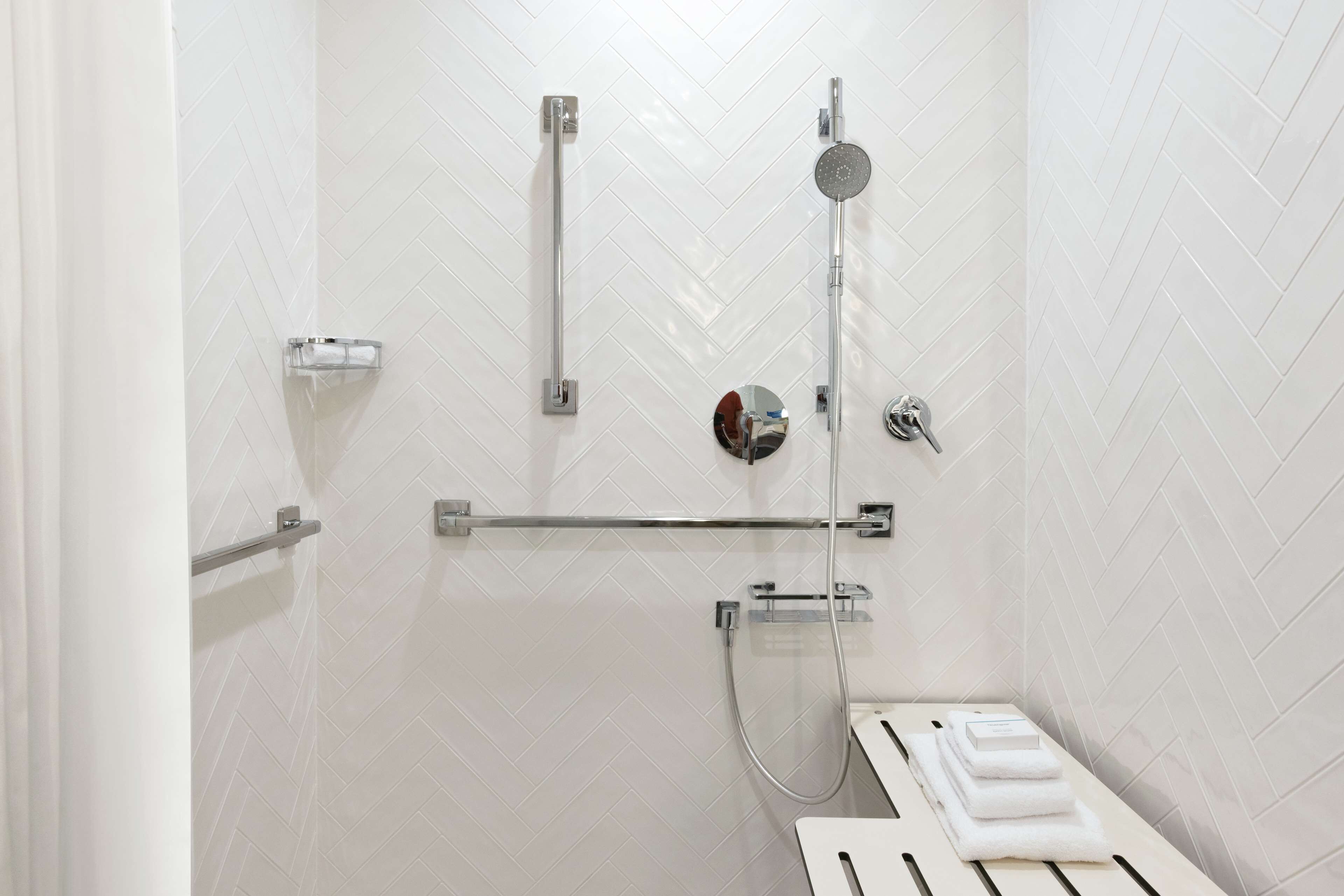 A modern bathroom with white herringbone tiles, a glass shower enclosure, a handheld showerhead, and a towel rack.