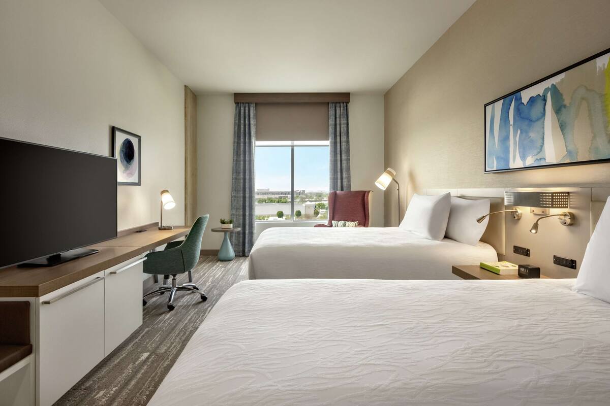 A hotel room with two beds, a desk with a TV, a chair, a lamp, and a painting on the wall.