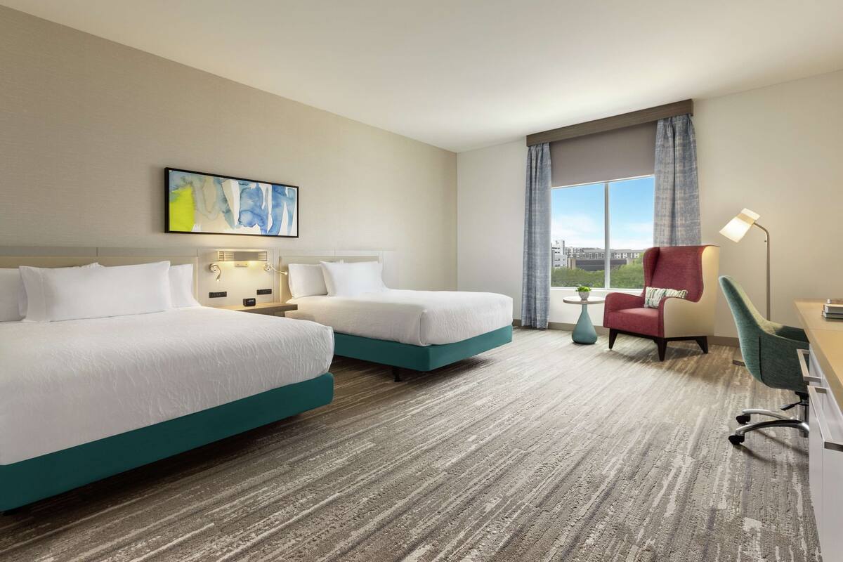A hotel room with two beds, a desk, a chair, a lamp, and a view of the city.