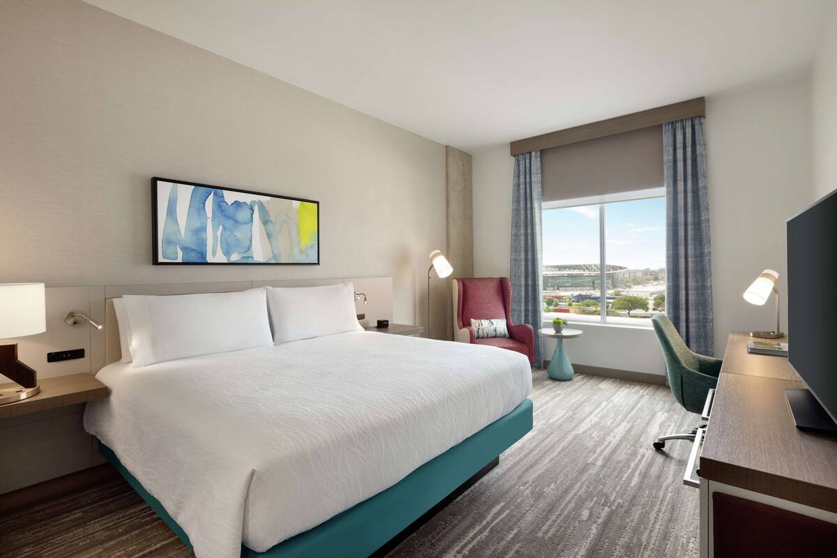 A hotel room with a large bed, a desk with a computer, a chair, a window with a view, and a painting on the wall.