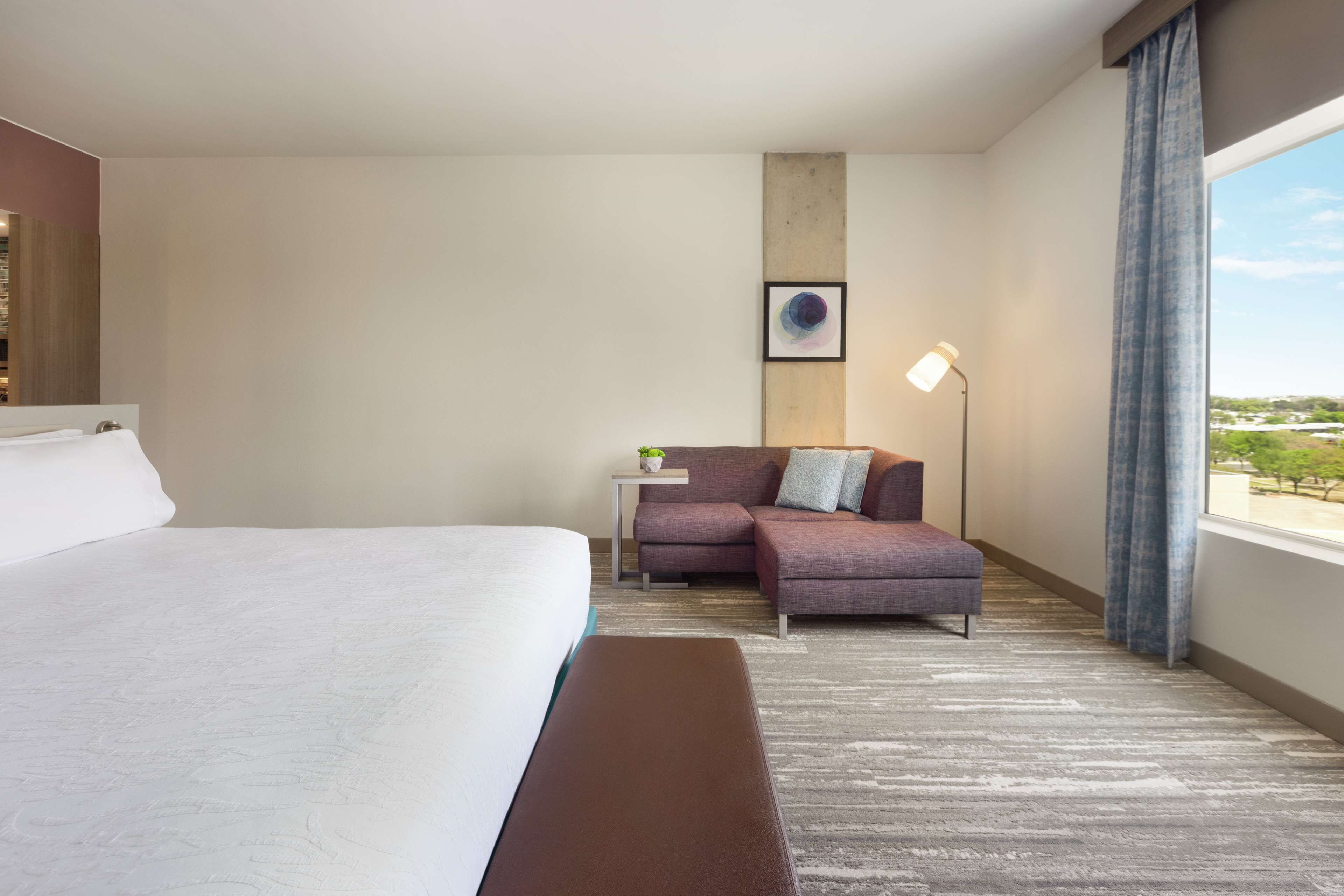 Quarto de hotel com uma cama grande, um sofá, uma mesinha com abajur e vista para o exterior.