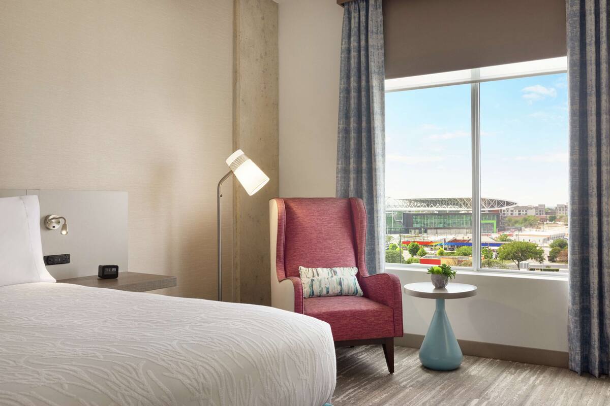 A hotel room with a large bed, a red armchair, a small round table with a plant, a floor lamp, and a window with a city view.