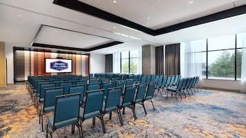 Un salón de conferencias con filas de sillas azules, una pantalla grande que muestra 'Hampton Inn & Suites' y ventanas amplias con vistas a árboles.