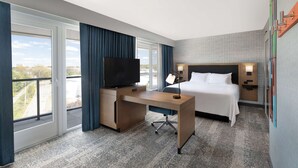 A hotel room with a bed, a desk with a TV, a large window with curtains, and a city view.