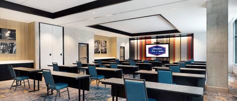 Un salón de conferencias con varias mesas y sillas, una pantalla grande mostrando el logotipo de Hampton Inn, y una decoración de diseño moderno.