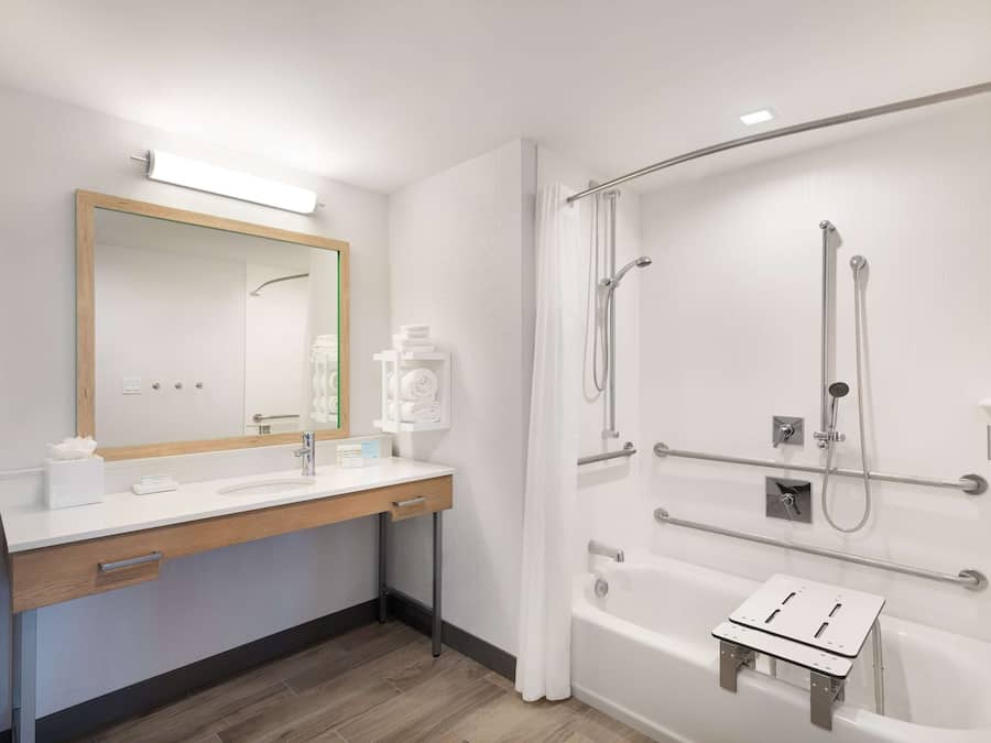 A modern bathroom with a wooden vanity, a large mirror, a bathtub with a showerhead, and a towel rack.