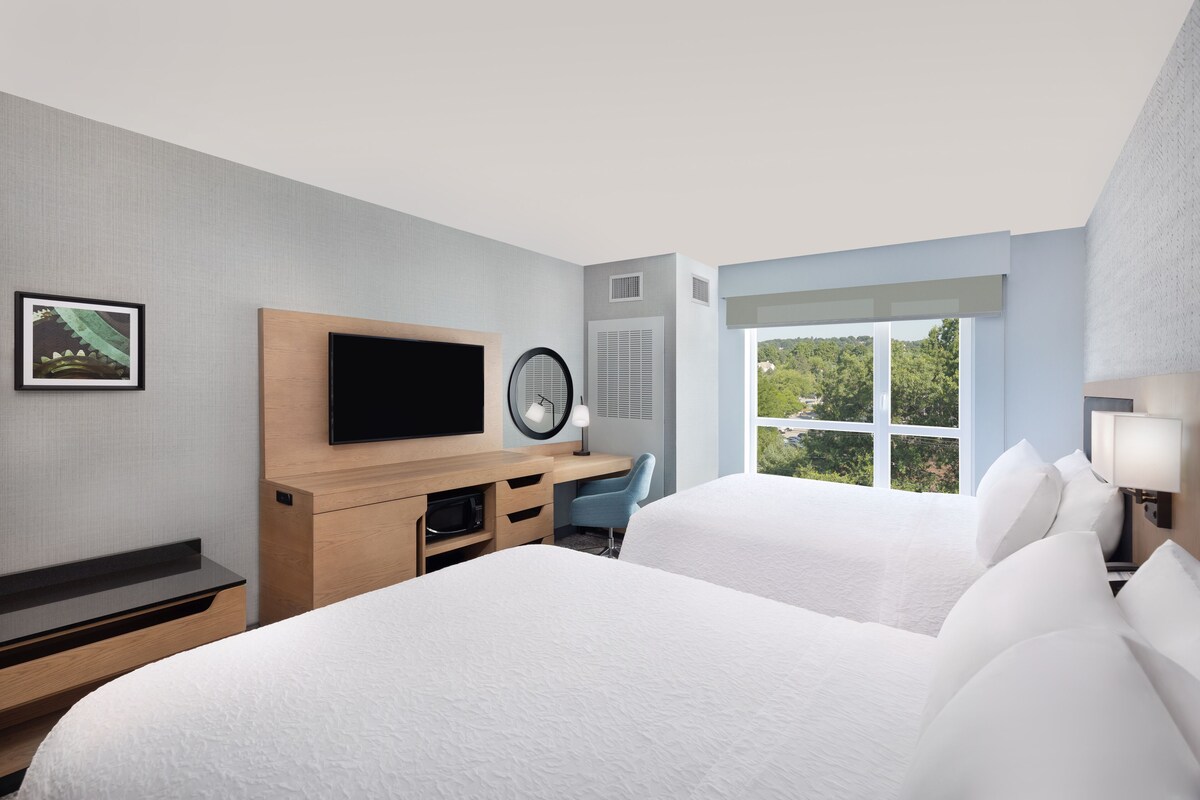 A hotel room with two beds, a TV, a desk with a chair, a window with a view of greenery, and a framed picture on the wall.