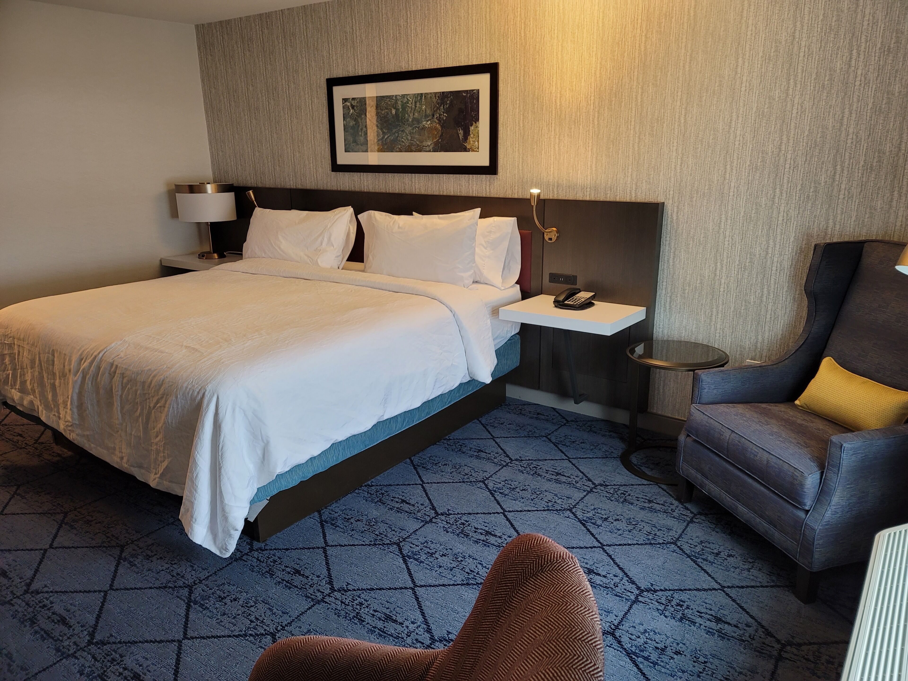 A hotel room with a bed, bedside table, lamp, chair, and a framed picture on the wall.