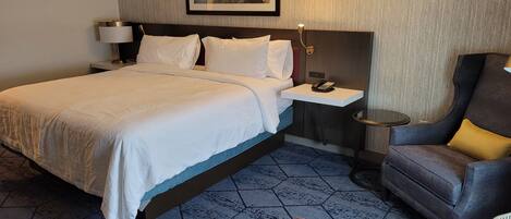 A hotel room with a bed, bedside table, lamp, chair, and a framed picture on the wall.