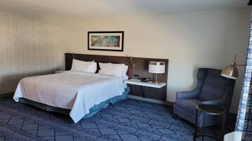 A hotel room with a large bed, a bedside table with a lamp, a chair, and a small table.