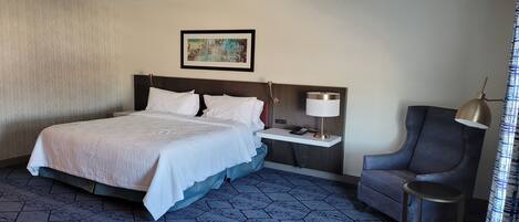 Una habitación de hotel con una cama grande, una mesita de noche con lámpara, una silla y una pequeña mesa.