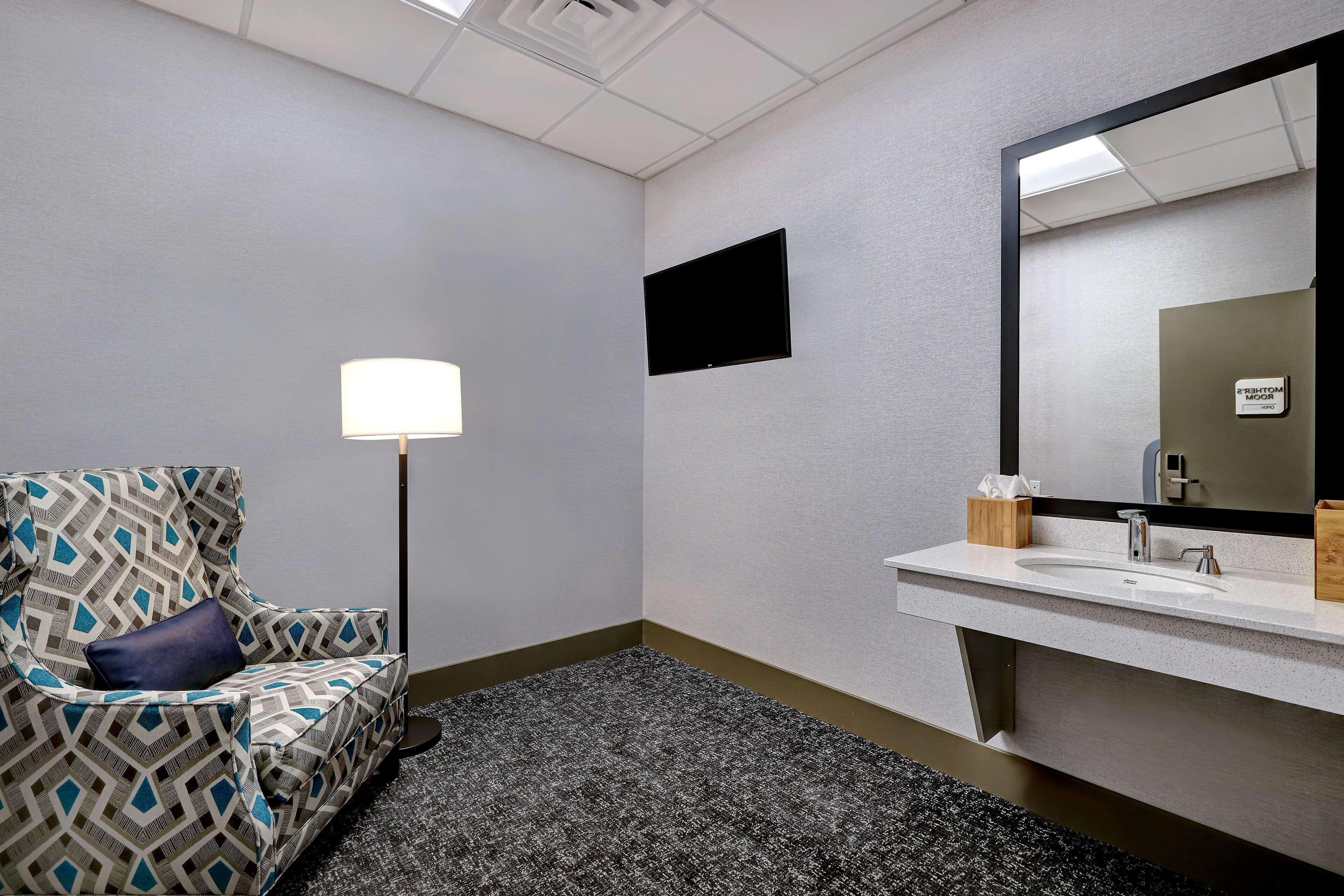Ein Hotelzimmer mit einem gemusterten Sessel, einer Nachttischlampe, einem Flachbildfernseher und einem modernen Waschbecken mit Spiegel.