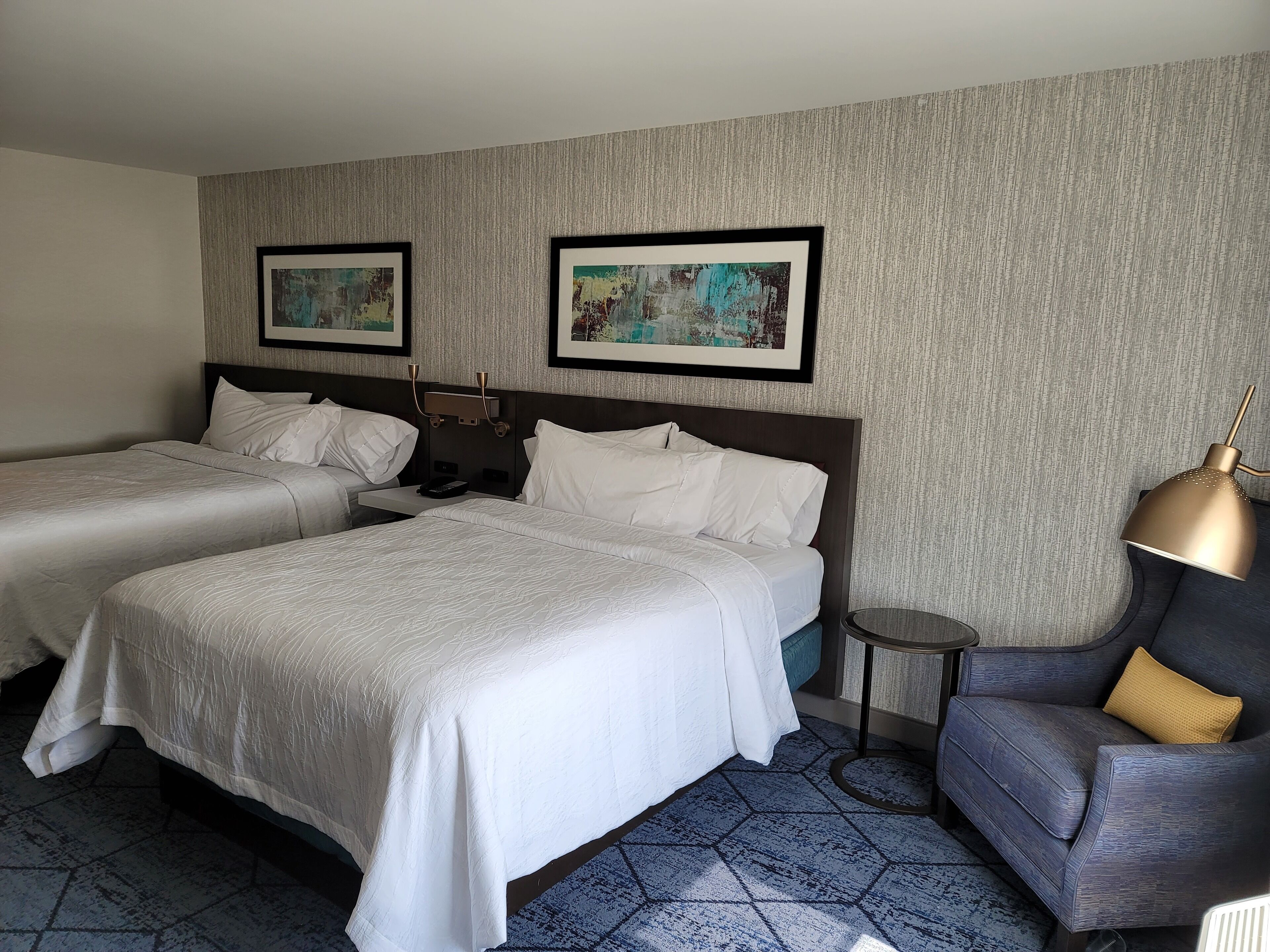 A hotel room with two beds, a small table, a chair, and two framed pictures on the wall.