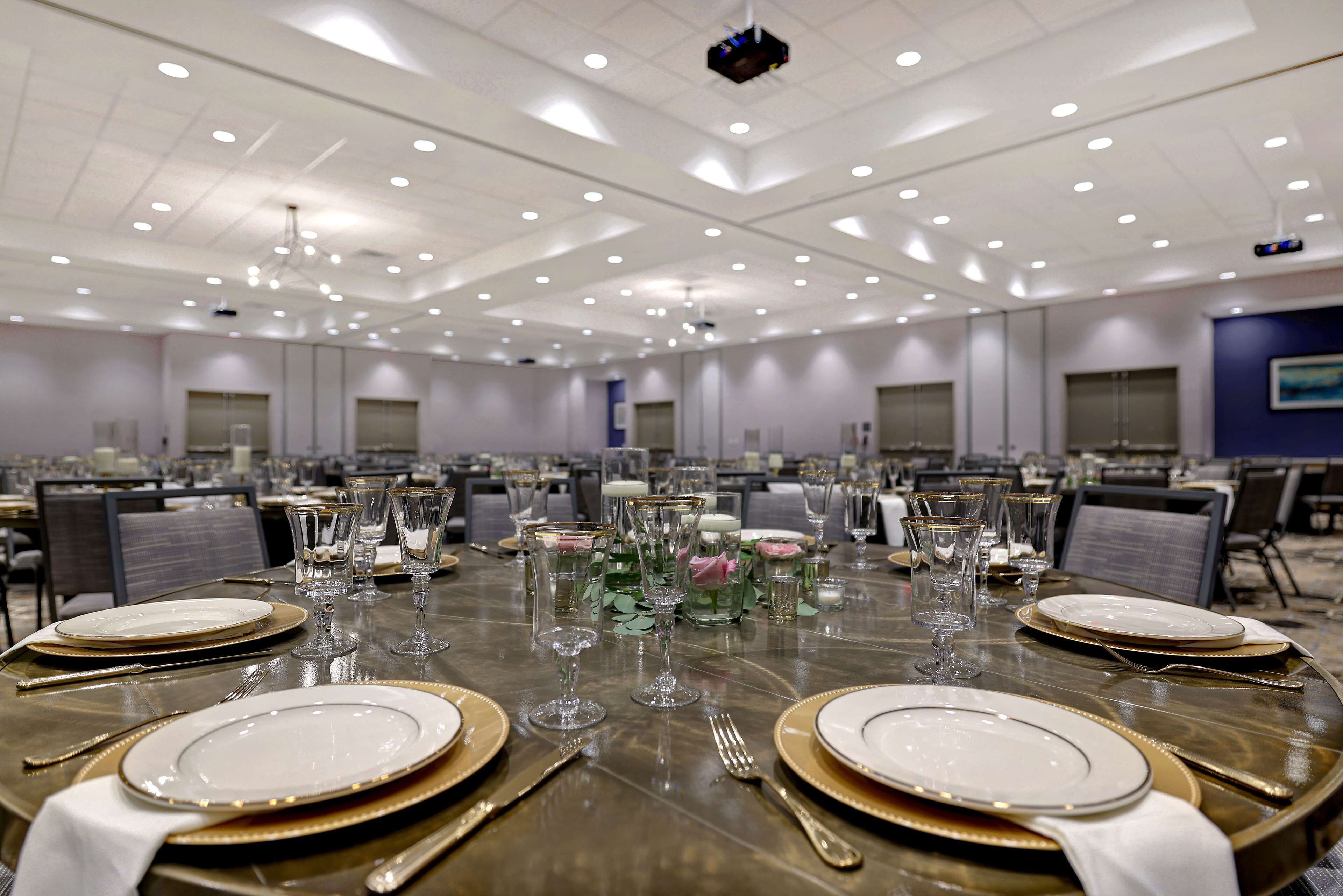 A large banquet hall with multiple round tables set for dining, equipped with gold-rimmed plates, silverware, and clear glassware.