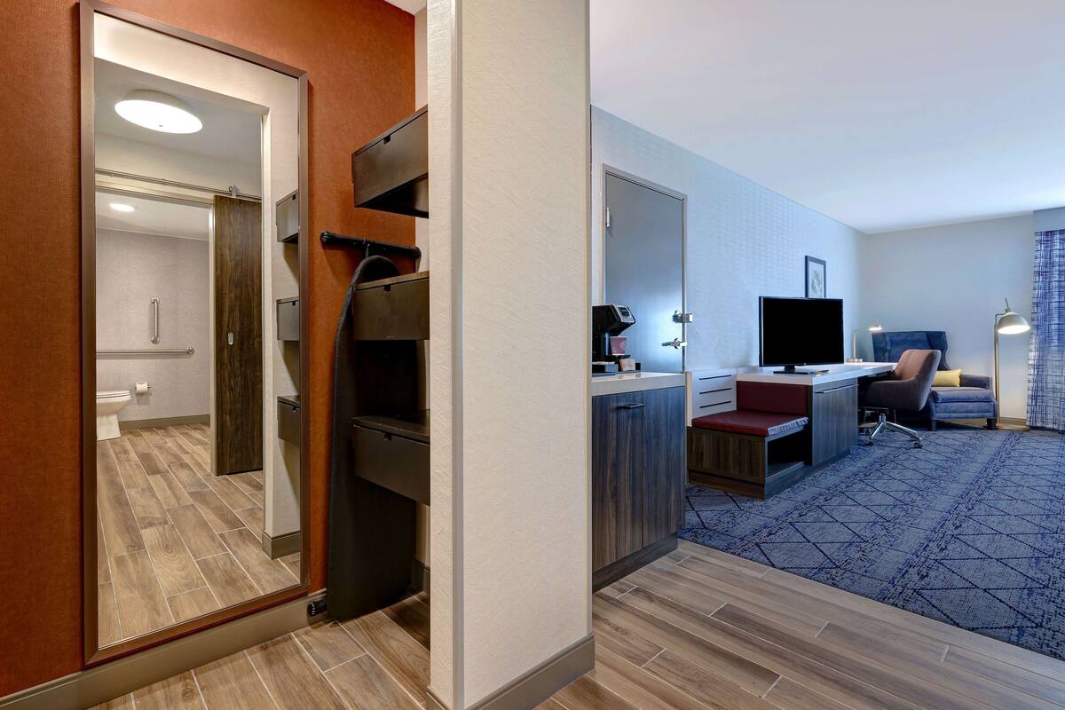 Ein Hotelzimmer mit Spiegel, Schreibtisch inklusive Computer, Stuhl, Fernseher und einem gemusterten Teppich.