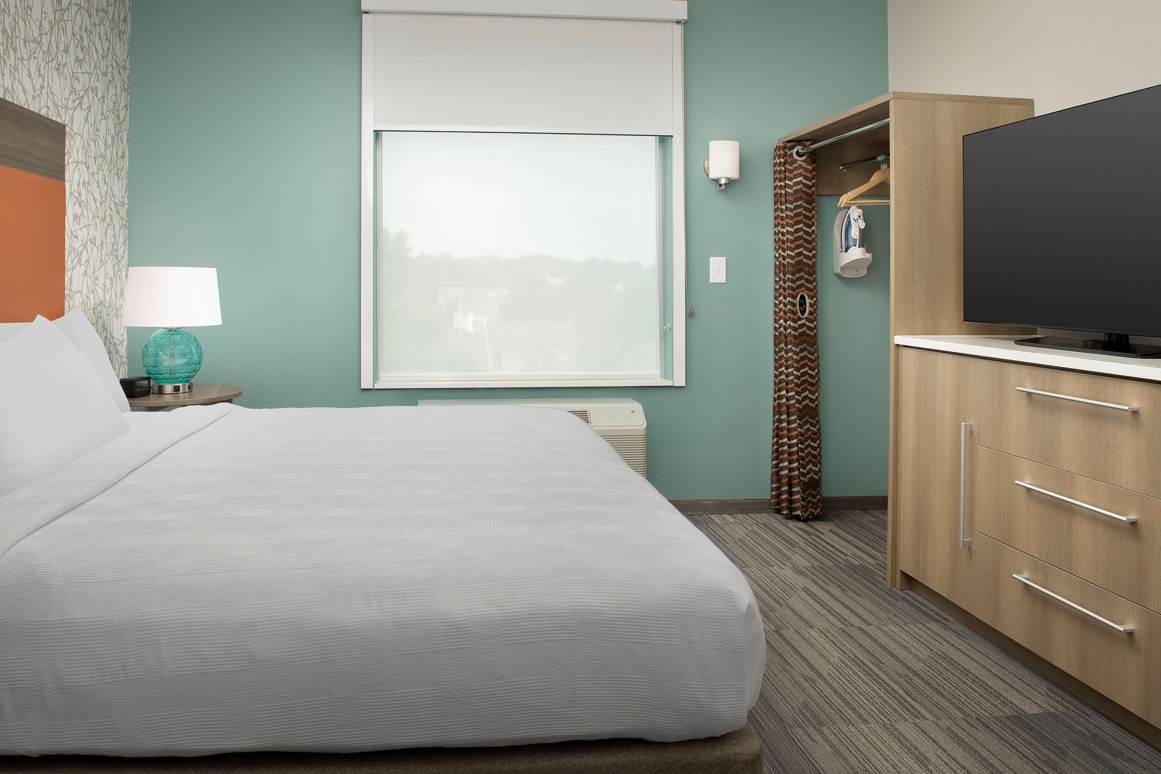 A hotel room with a bed, a TV, a wardrobe, and a window with a view of buildings.