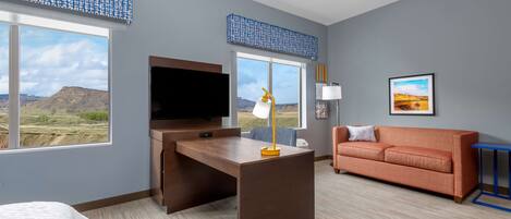 A hotel room with a desk, television, sofa, and large windows offering a view of the landscape.