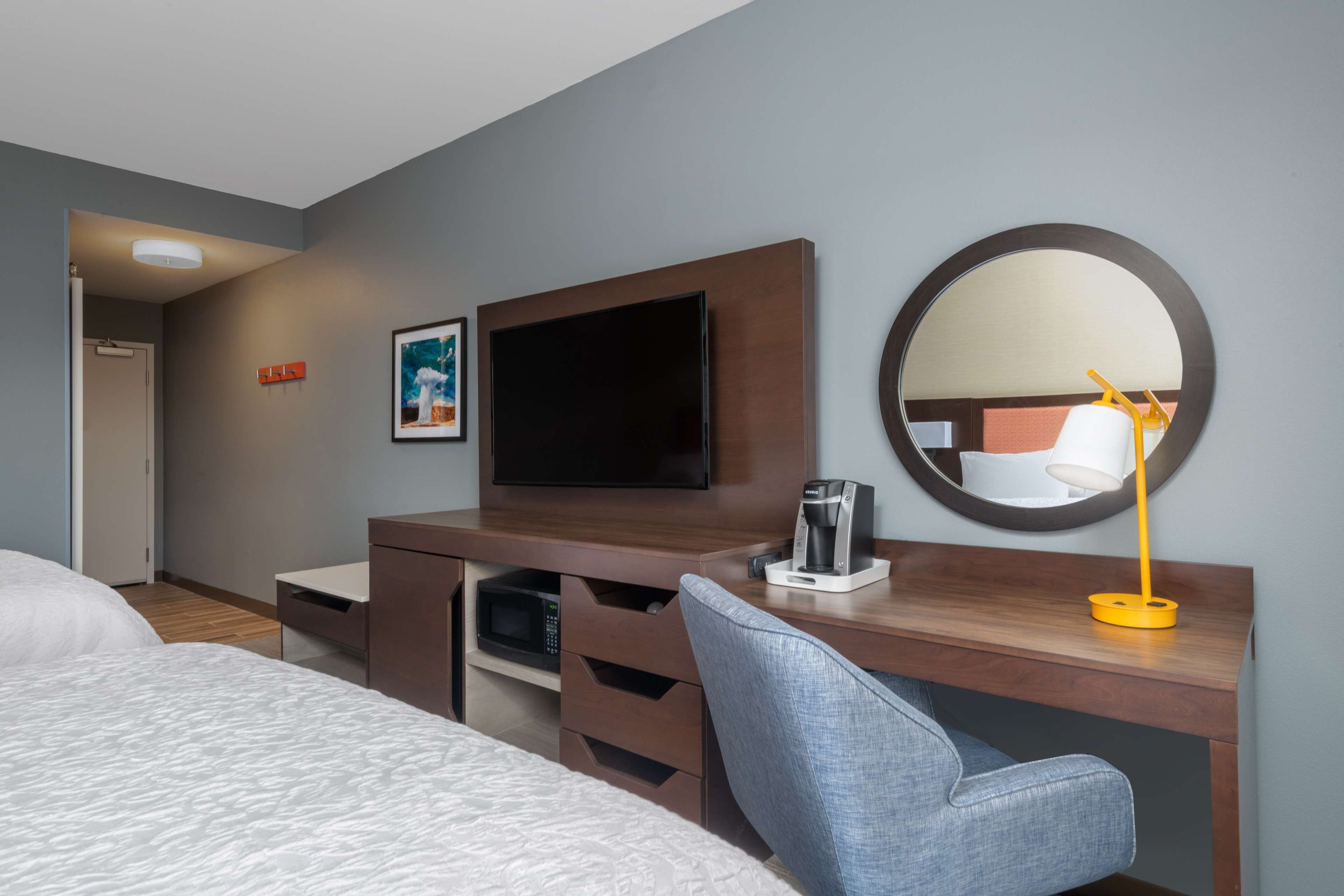 A hotel room with a bed, a desk with a flat-screen TV, a coffee maker, a mirror, and a framed picture on the wall.
