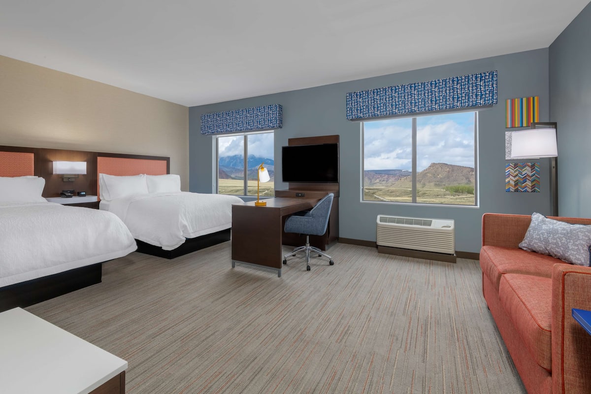 A hotel room with two beds, a desk, a TV, and a view of the mountains.