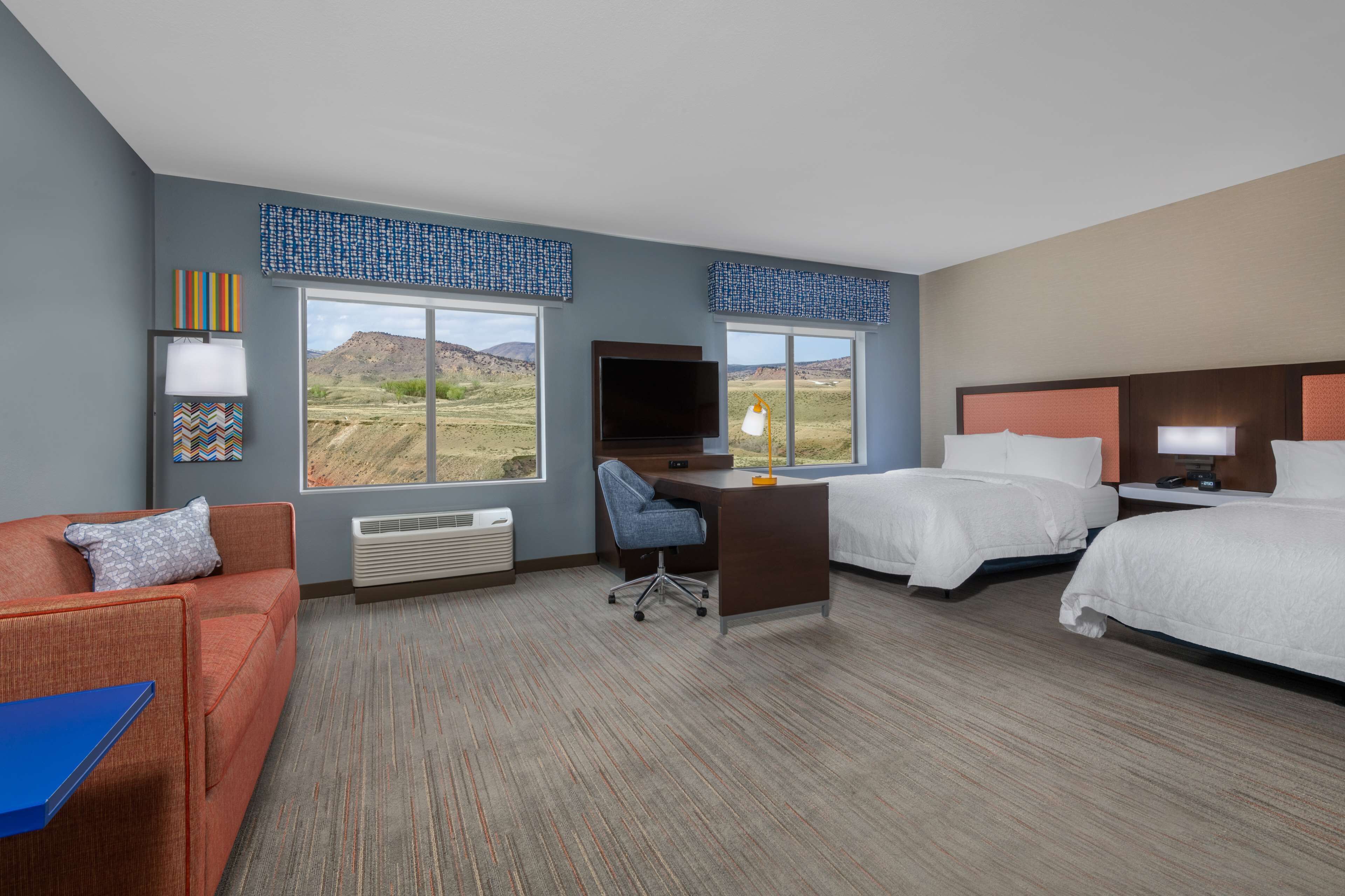 A hotel room with a desk, two beds, a sofa, and large windows offering a view of the desert landscape.
