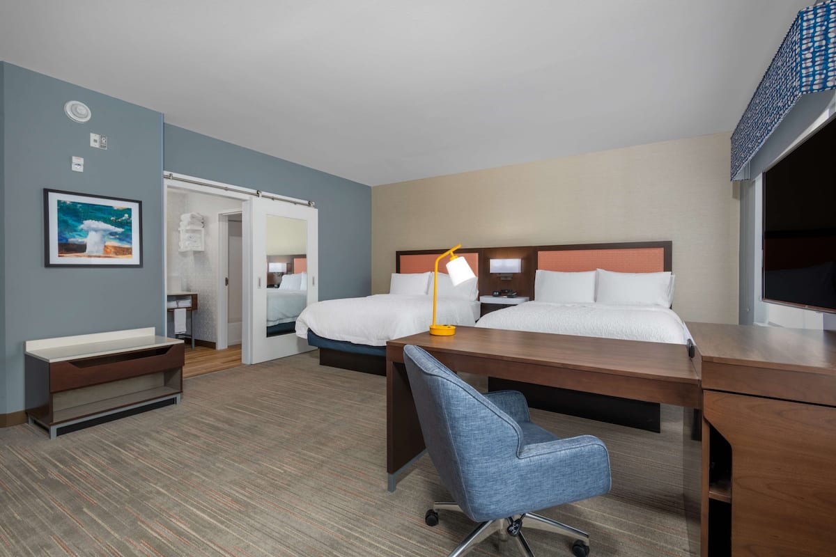 A hotel room with two beds, a desk, a chair, a desk lamp, a TV, and a painting on the wall.
