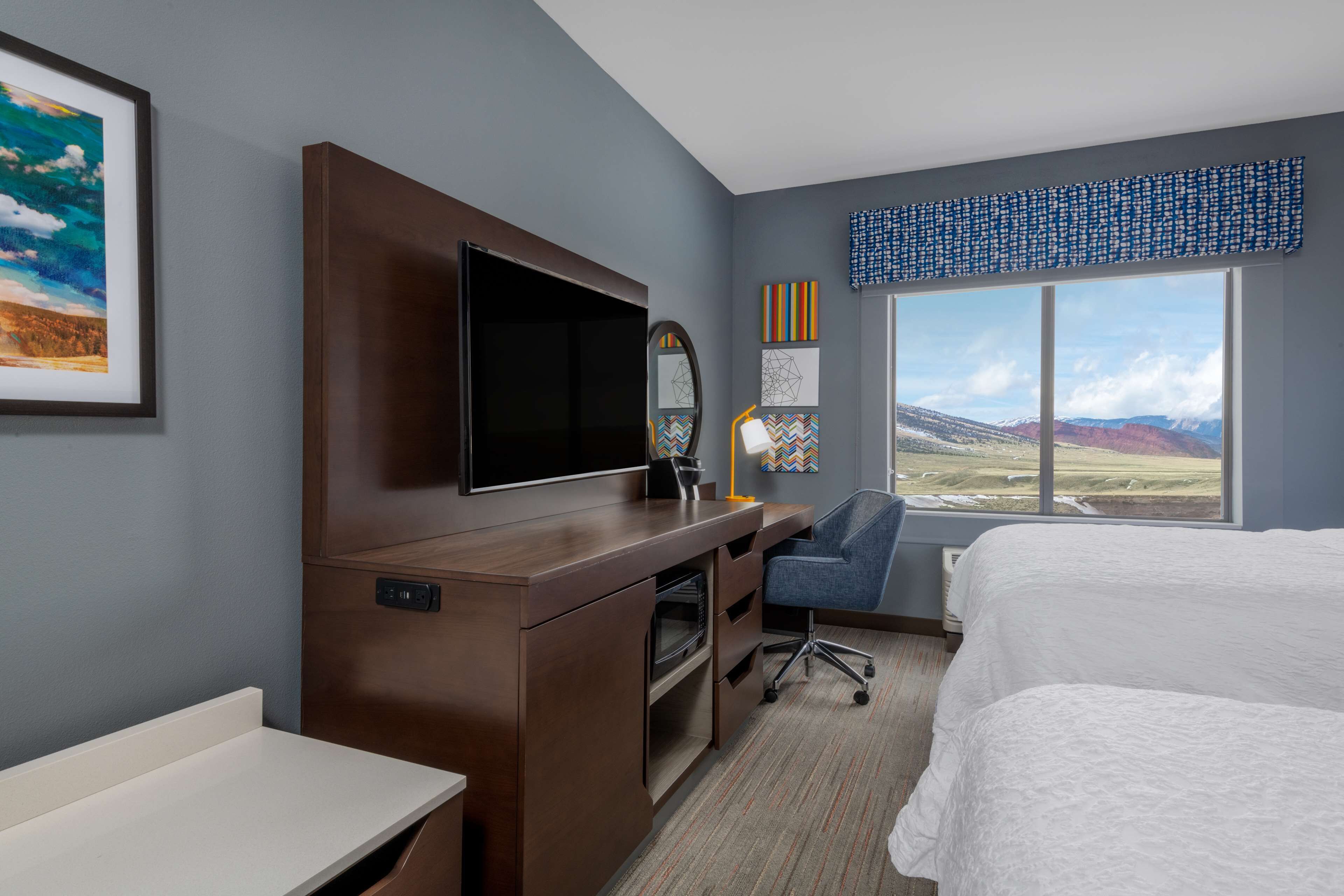 A hotel room with a bed, desk, TV, and a large window with a mountain view.