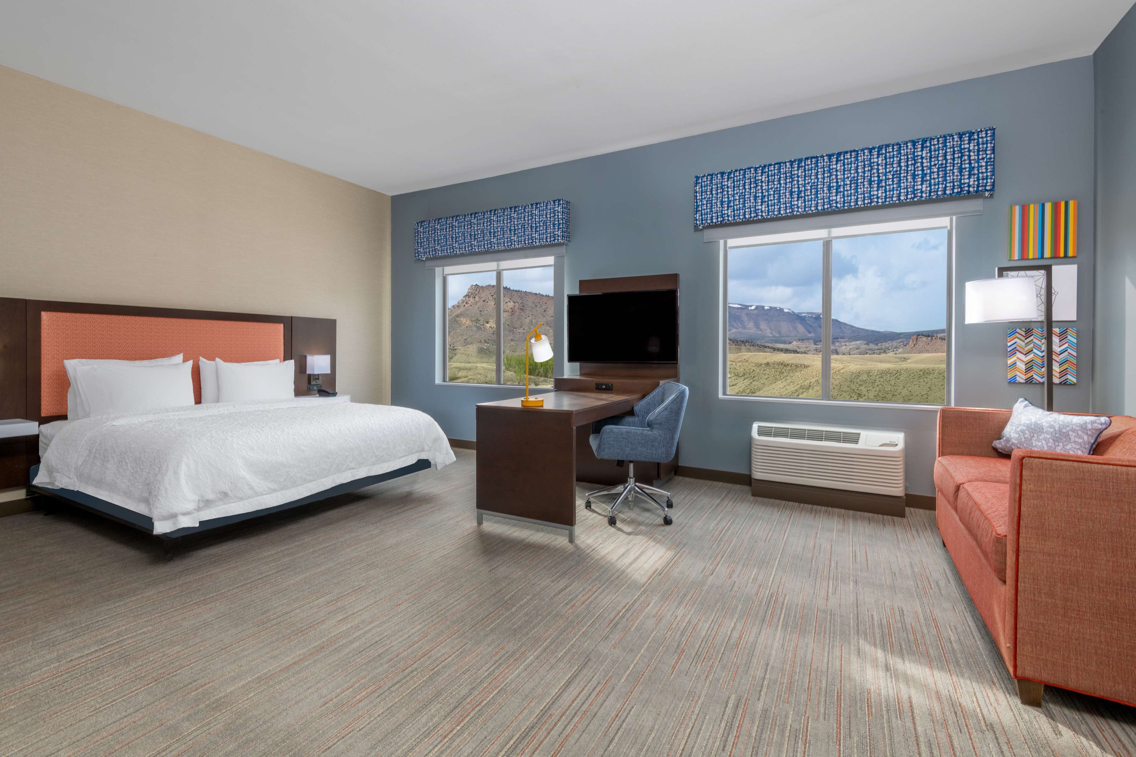 Une chambre d’hôtel équipée d’un lit, d’un bureau et d’une chaise, d’une télévision et de grandes fenêtres offrant une vue sur les montagnes.