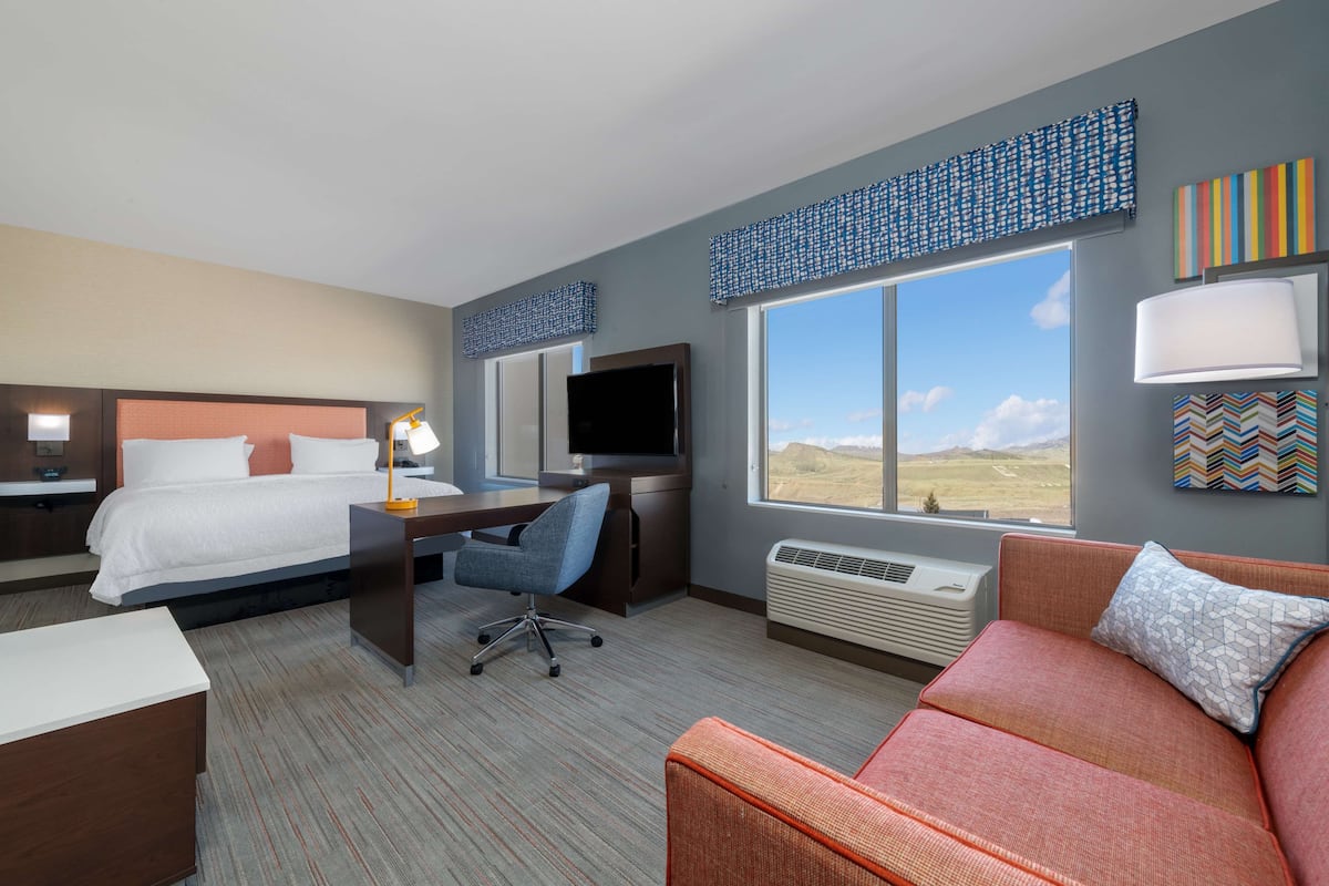 A hotel room with a bed, desk, chair, couch, and large windows offering a view of the outdoors.