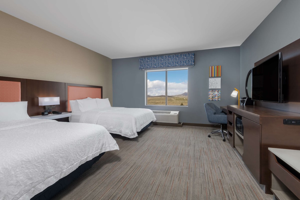 A hotel room with two beds, a desk, a chair, a TV, and a large window with a view of the outdoors.