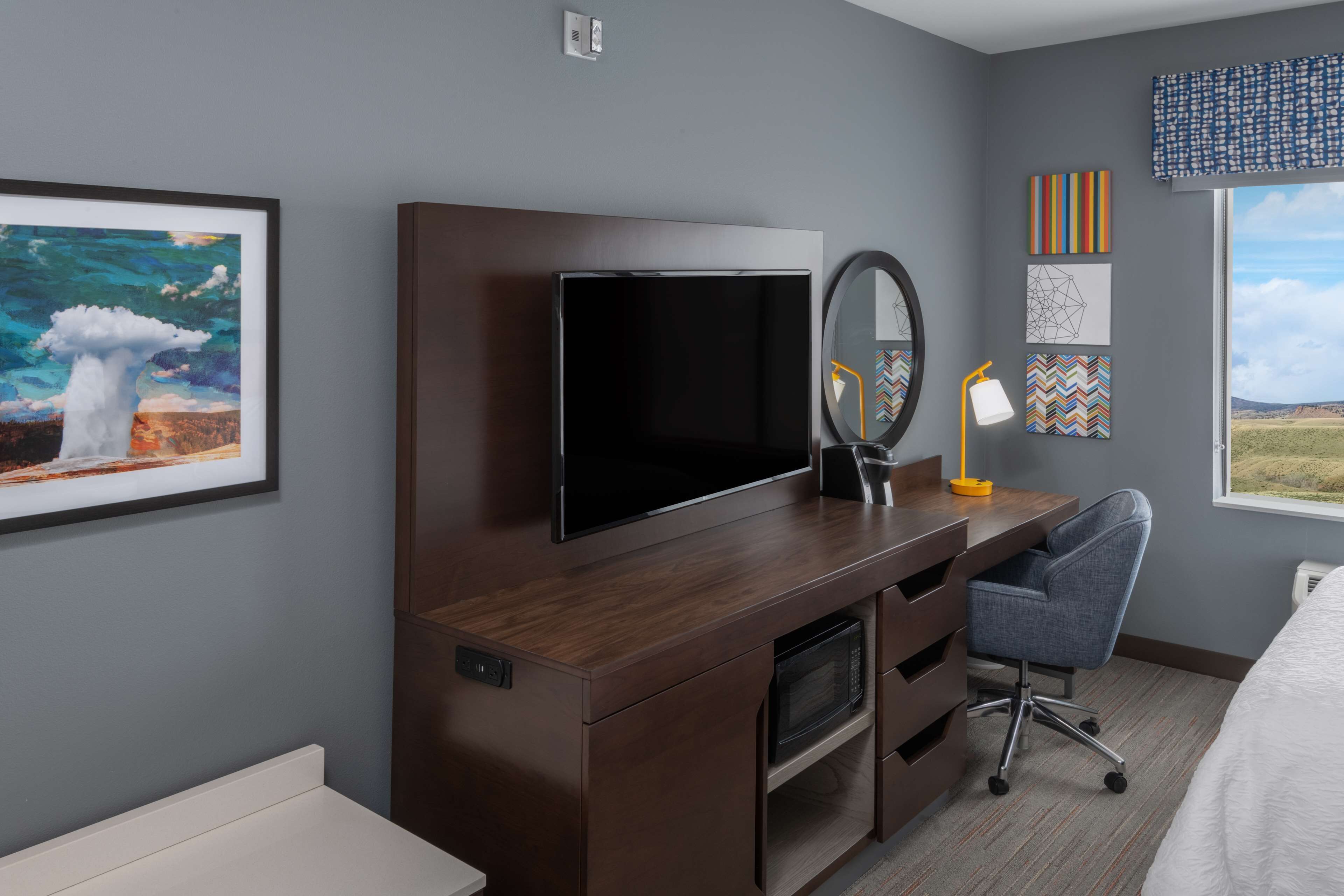 Habitación de hotel con un escritorio de madera, televisor de pantalla plana, espejo, lámpara de escritorio, silla y obras de arte en las paredes.
