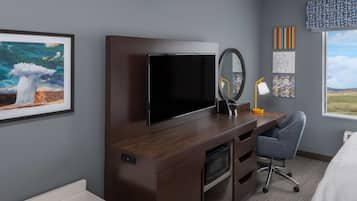Habitación de hotel con un escritorio de madera, televisor de pantalla plana, espejo, lámpara de escritorio, silla y obras de arte en las paredes.
