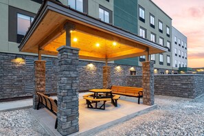 A covered outdoor seating area with stone pillars and a wooden roof, adjacent to a building with multiple windows. - Hampton Inn & Suites Cody (Cody)