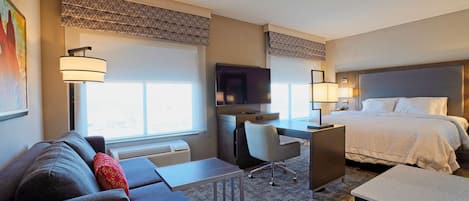 A hotel room with a bed, a desk, a chair, a sofa, a TV, and a window with blinds.