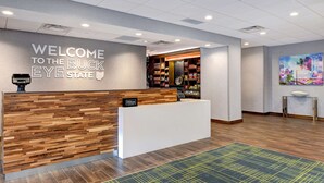 A hotel reception area with a wooden reception desk, a "Welcome to the Buck Eye State" sign, a Hilton logo, and a cityscape painting. - Hampton Inn & Suites Canal Winchester Columbus (Canal Winchester)