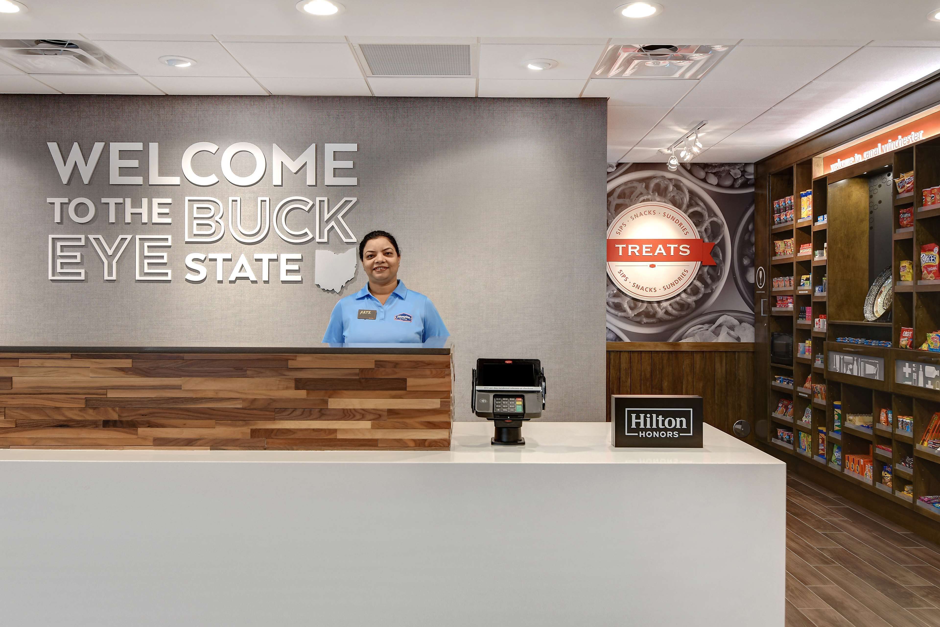 A reception desk with a "WELCOME TO THE BUCK EYE STATE" sign, a "Hilton Honors" plaque, and a "TREATS" sign indicating a snack area.