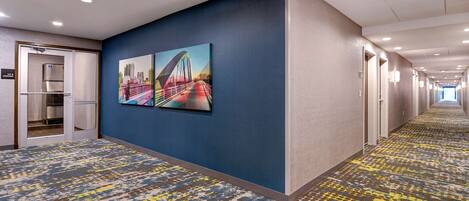 A hotel hallway with a patterned carpet, a refrigerator in the corner, and a large cityscape painting on the wall.