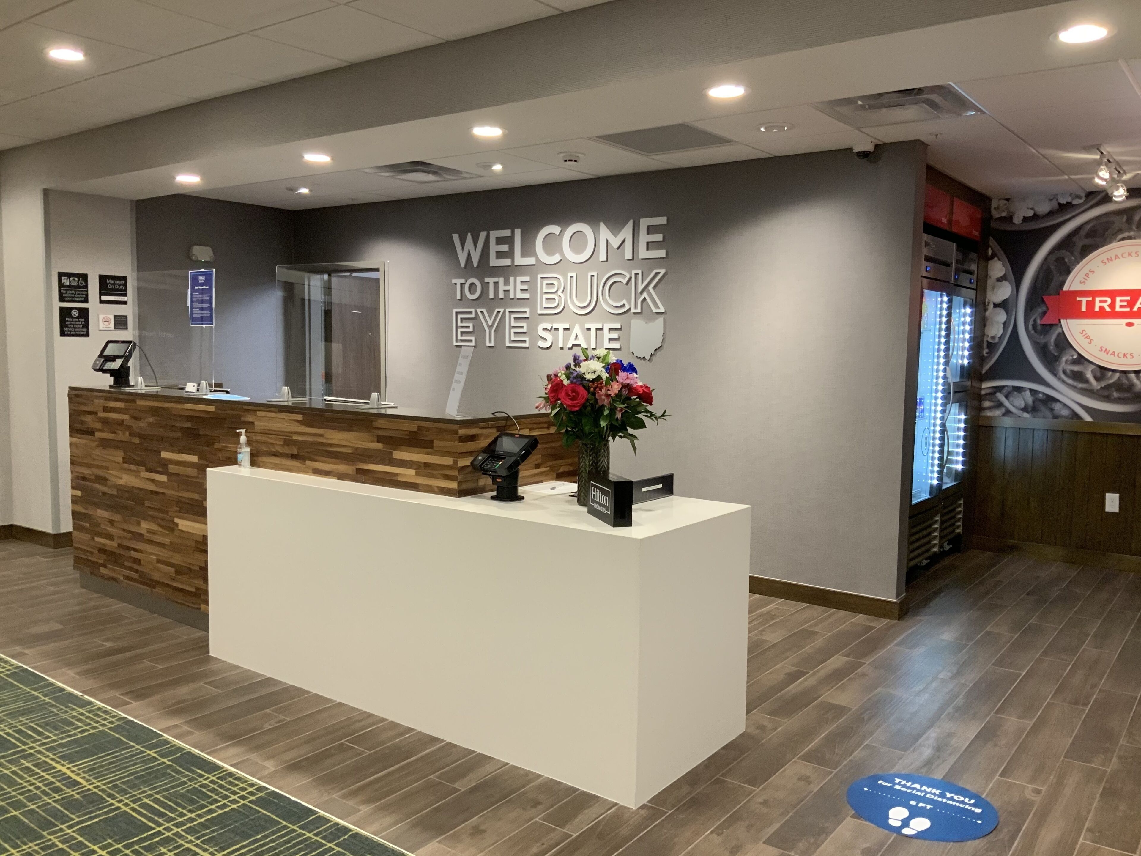 A reception area with a 'Welcome to the Buck Eye State' sign, a reception desk, a floral arrangement, and a 'Thank You for Social Distancing' mat.