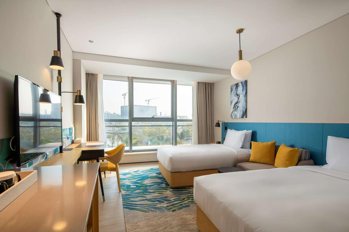 A hotel room with two beds, a desk, a TV, and a large window with a view of the city.