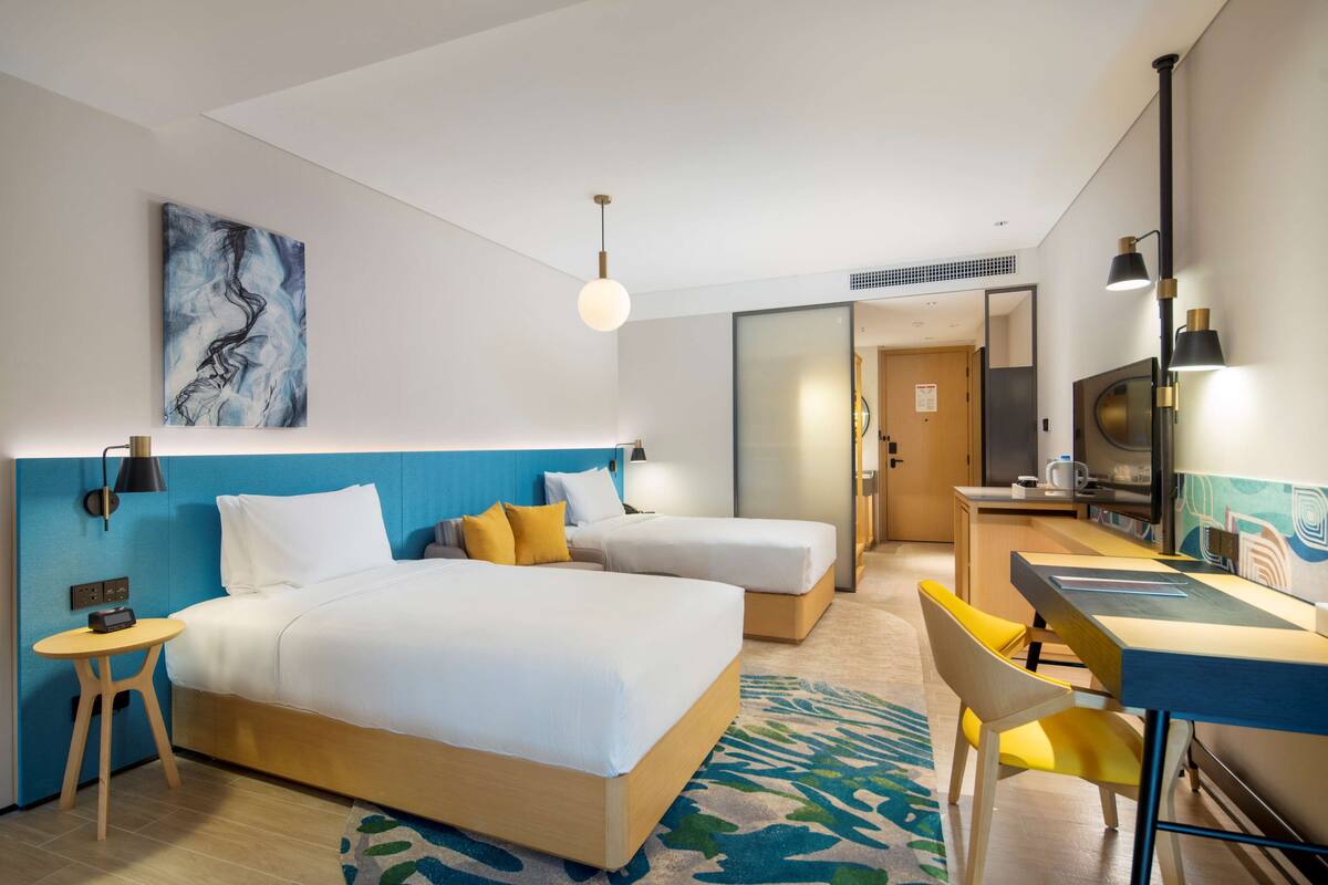 A hotel room with a large bed, a smaller bed, a desk, a chair, a table, a lamp, a painting, and a door leading to a bathroom.