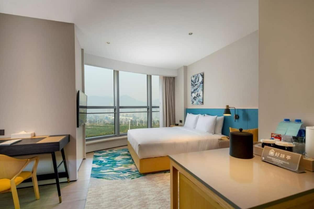 A hotel room with a bed, desk, chair, and a large window offering a view of the cityscape.