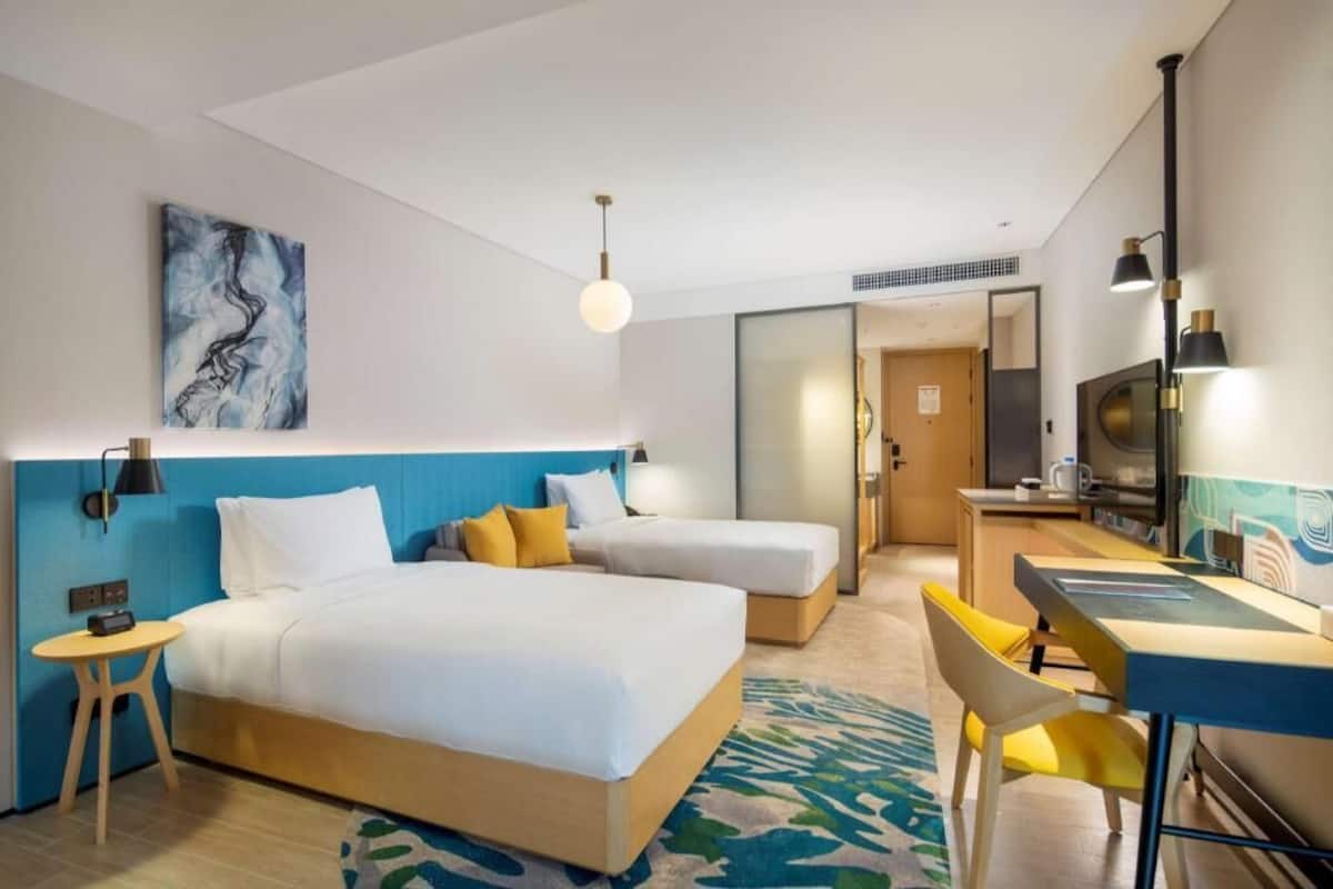 A hotel room with a bed, two bedside tables, a desk, a chair, a TV, and a painting on the wall.